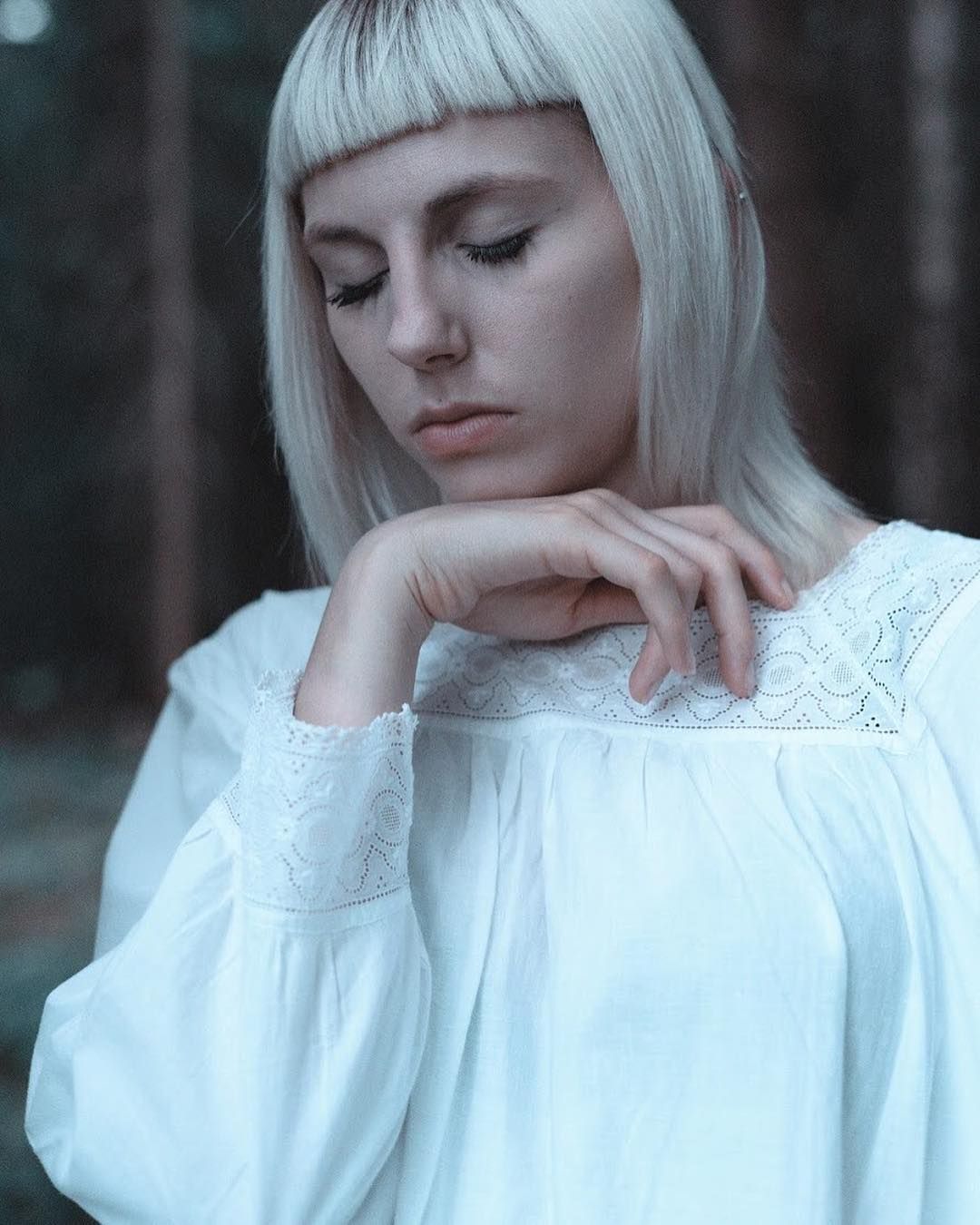 A woman in a white lace dress resting her chin on her hand, soft forest light behind her