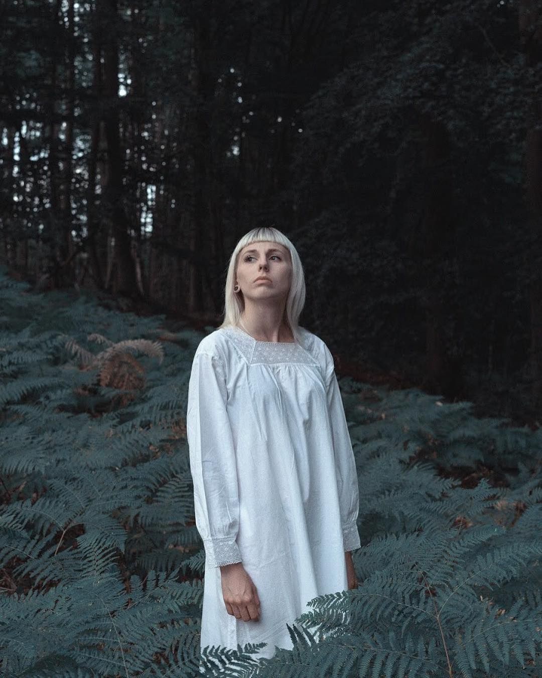 A woman in white standing among ferns, looking up into a darkening forest