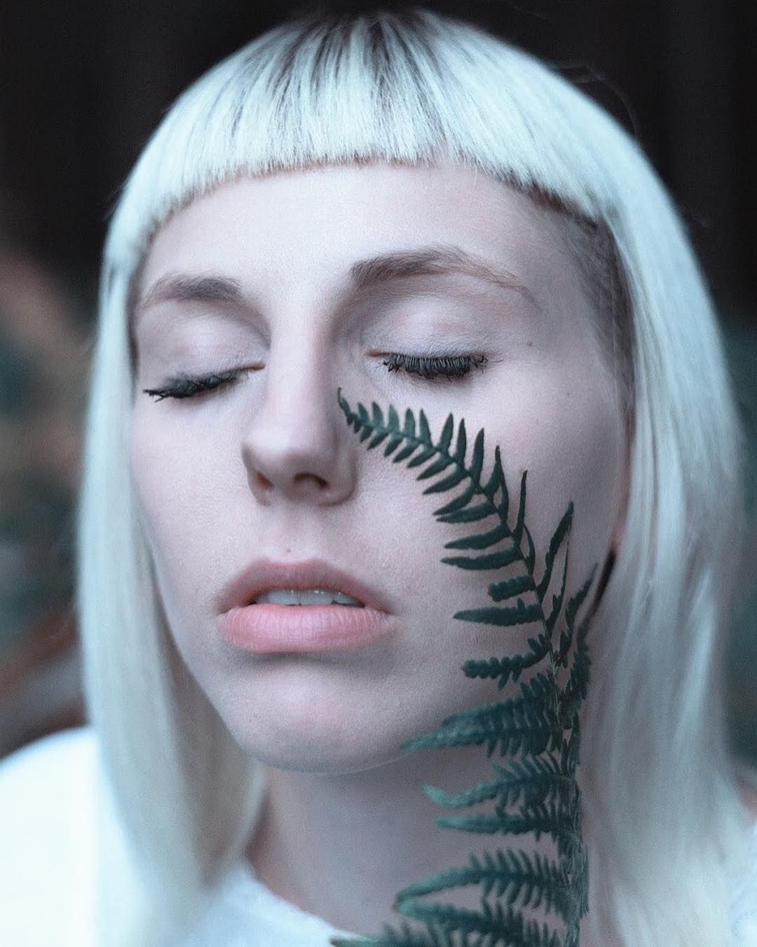 Close-up of a face with closed eyes, a fern leaf trailing across the cheek