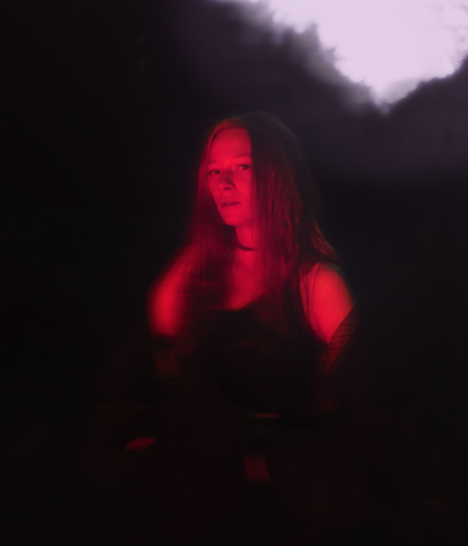 A portrait lit red against a wall of trees, eyes closed and head tilted back