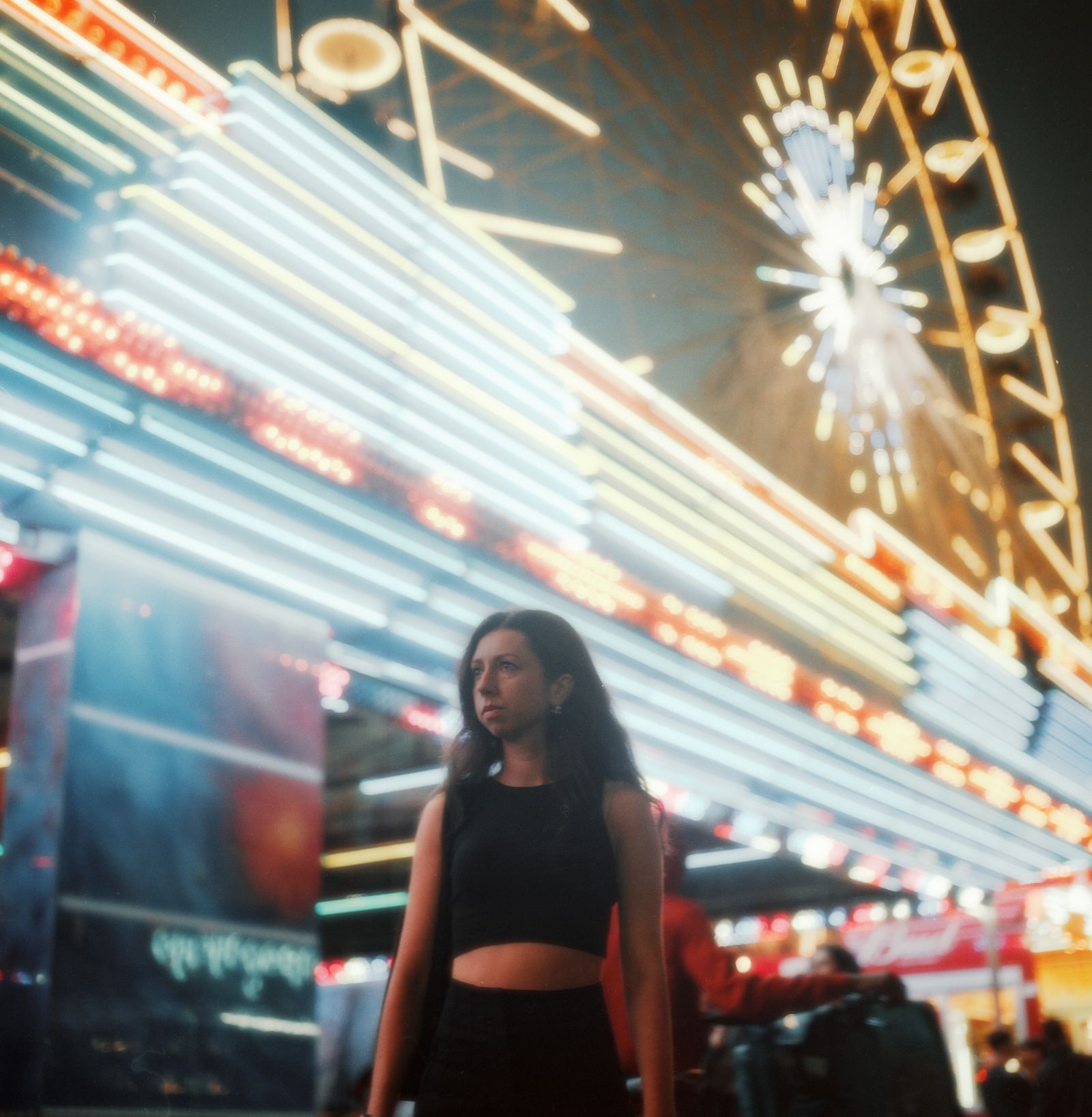 A woman standing before a brightly lit fairground ride, a ferris wheel glowing behind her