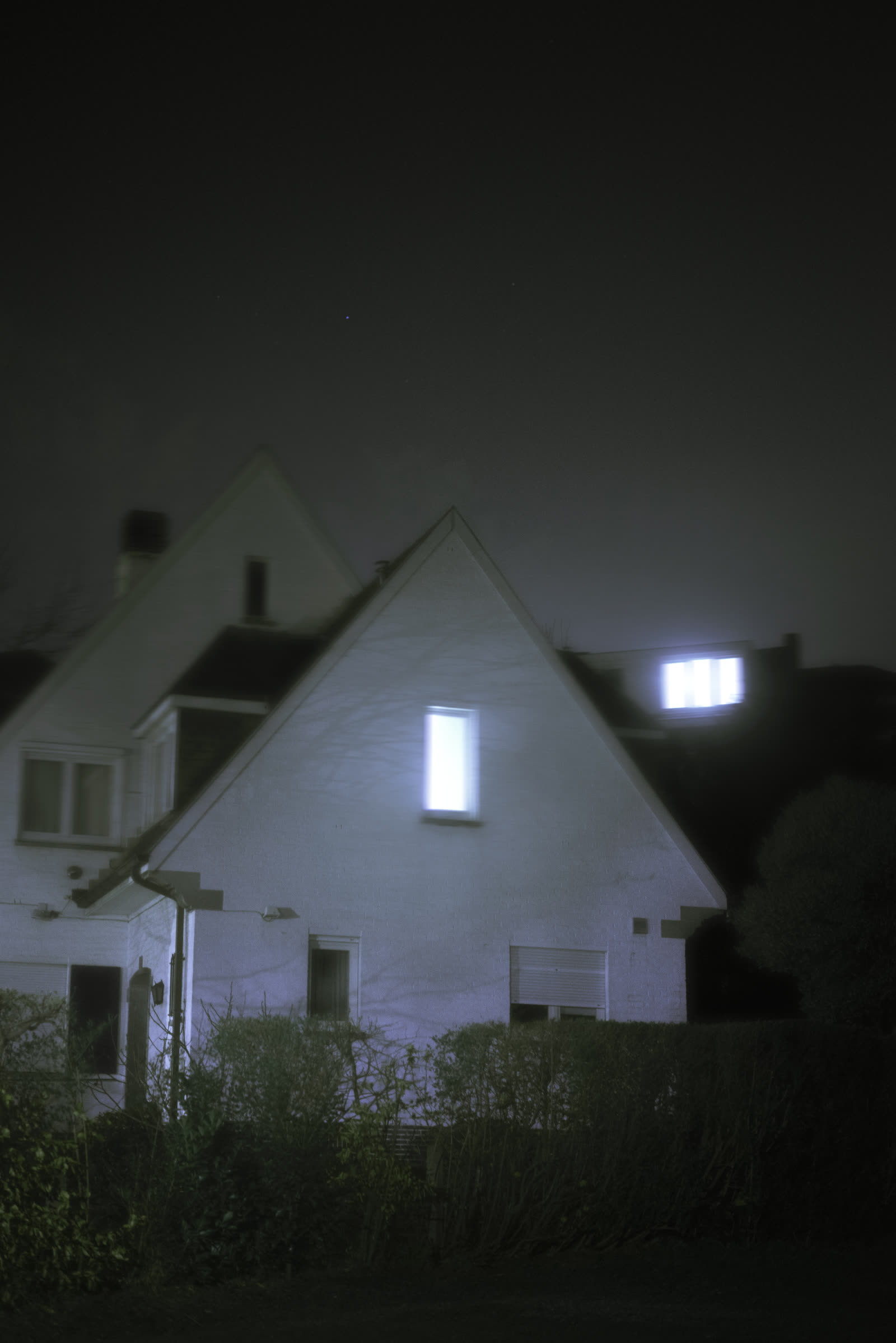 A house gable lit by a glowing attic window, another lit window on the roof beyond, hedges in the foreground