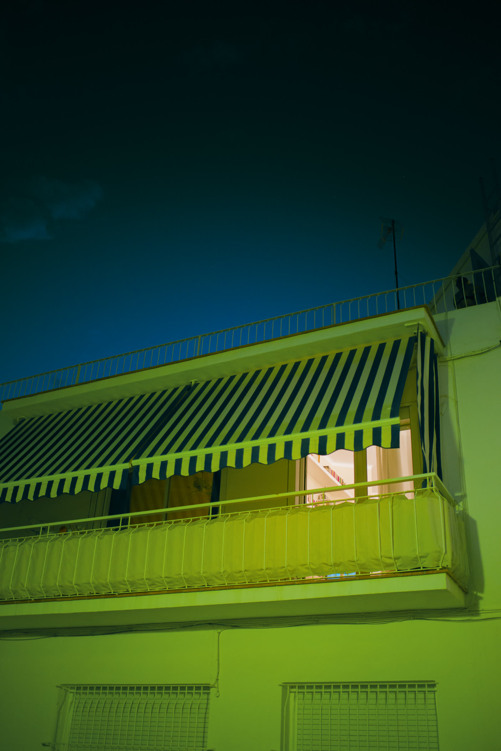A green-lit balcony with a striped awning, a warm window glowing behind it