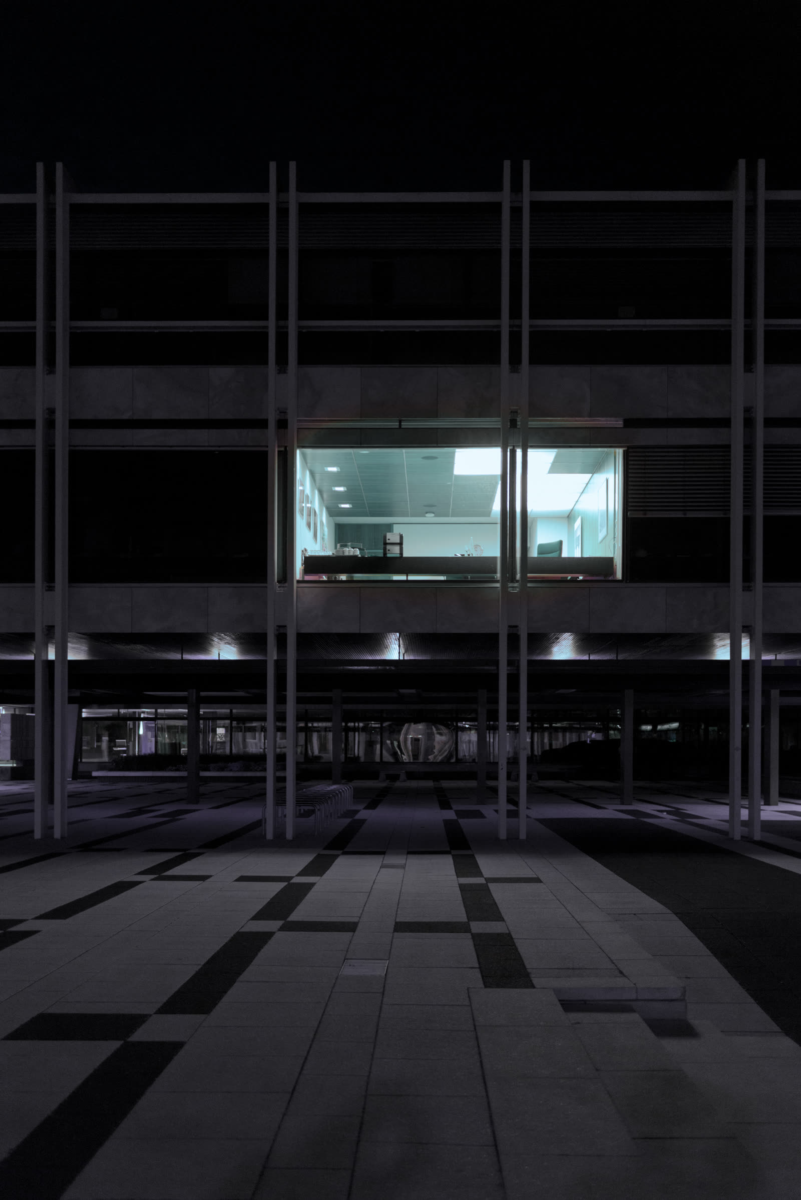 A modern courtyard at night — one office window lit behind rows of concrete pillars