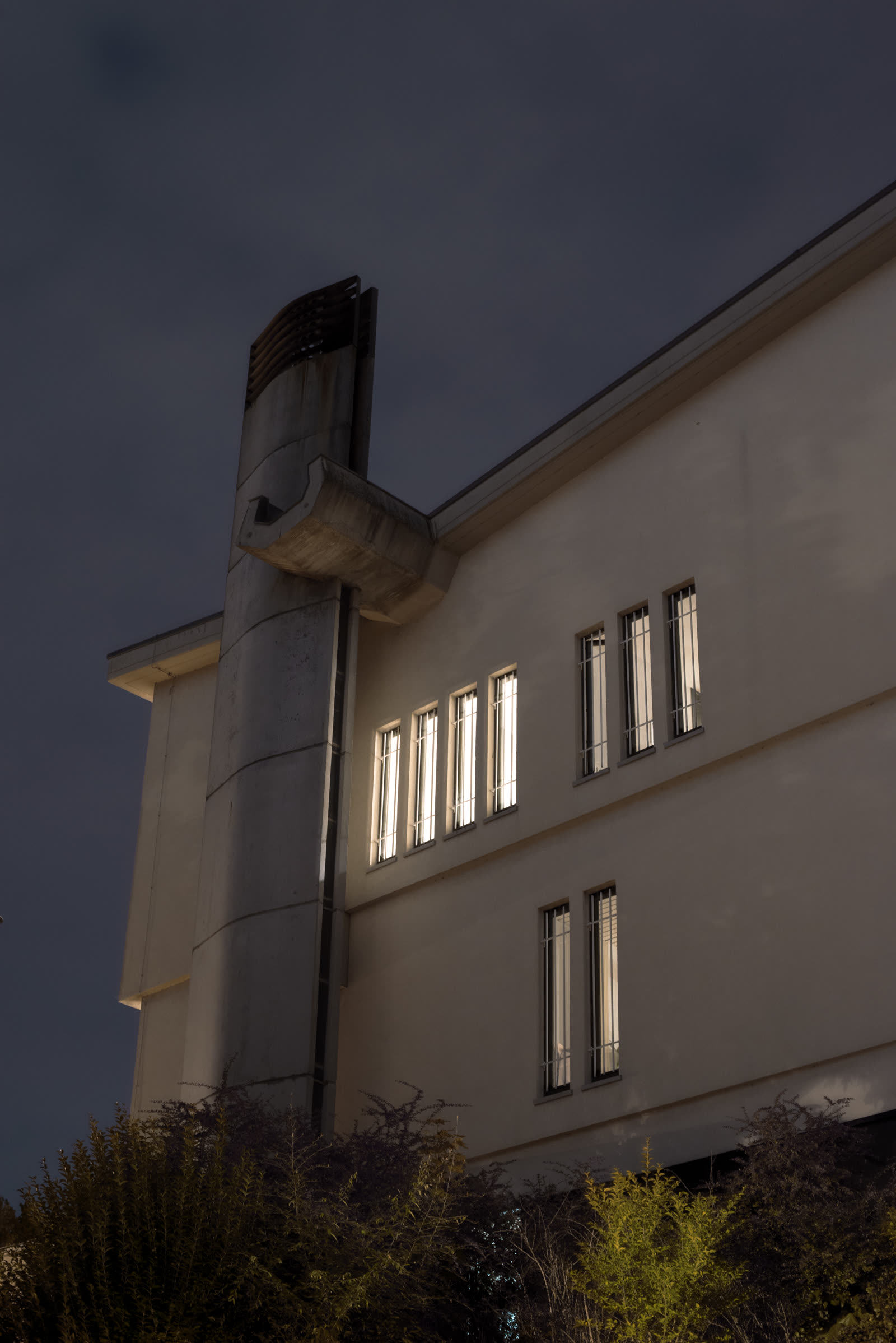 A building corner with a concrete duct, several windows glowing against the dusk