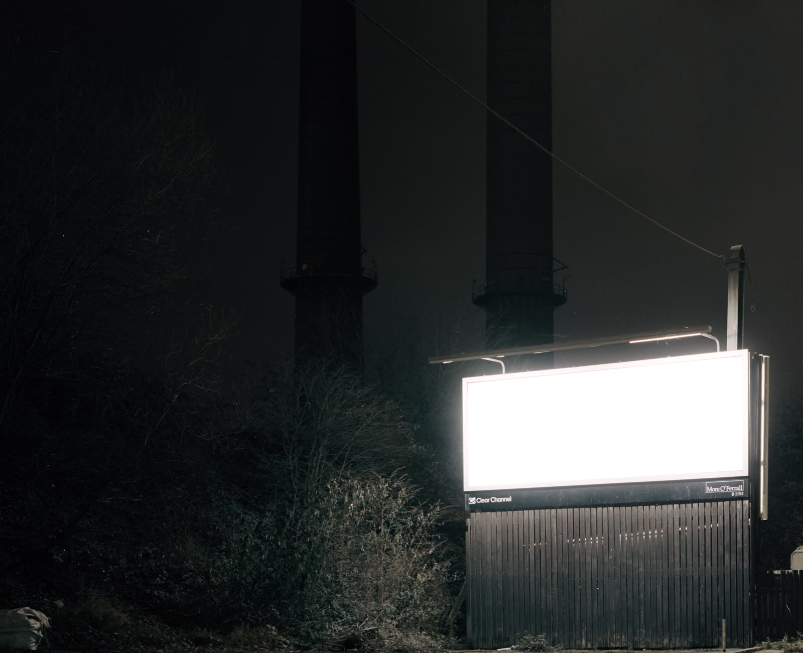 An empty illuminated billboard glowing white beside two dark industrial chimneys
