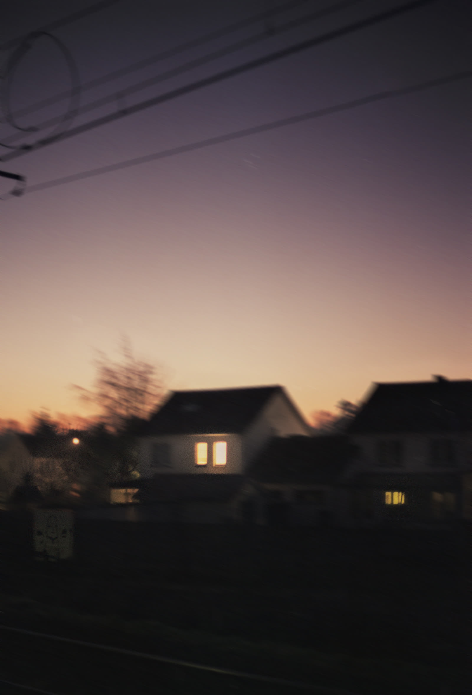 A pink dusk sky settling over rooftops, one window lit warm gold below