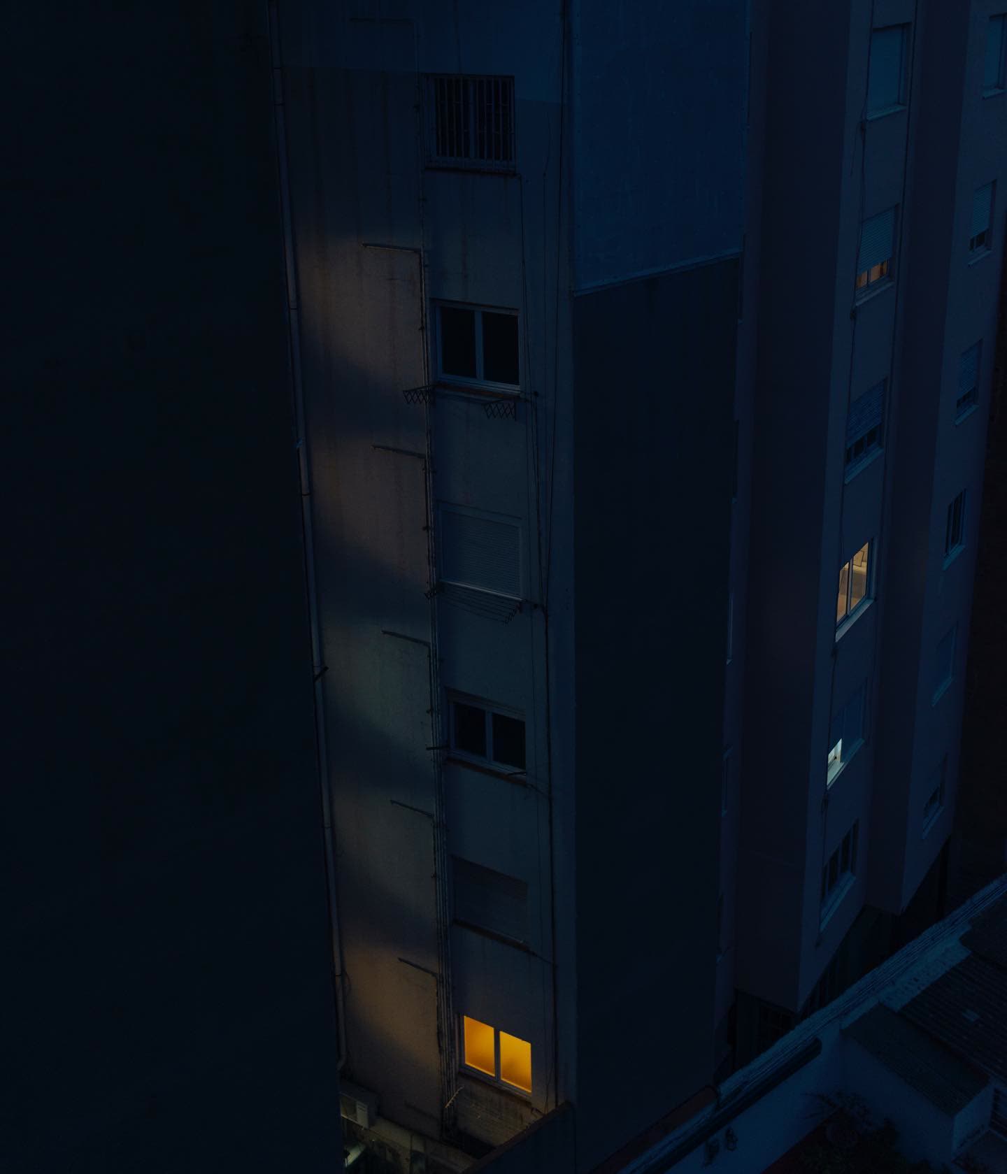 A tall apartment block, almost entirely dark — one window glowing orange