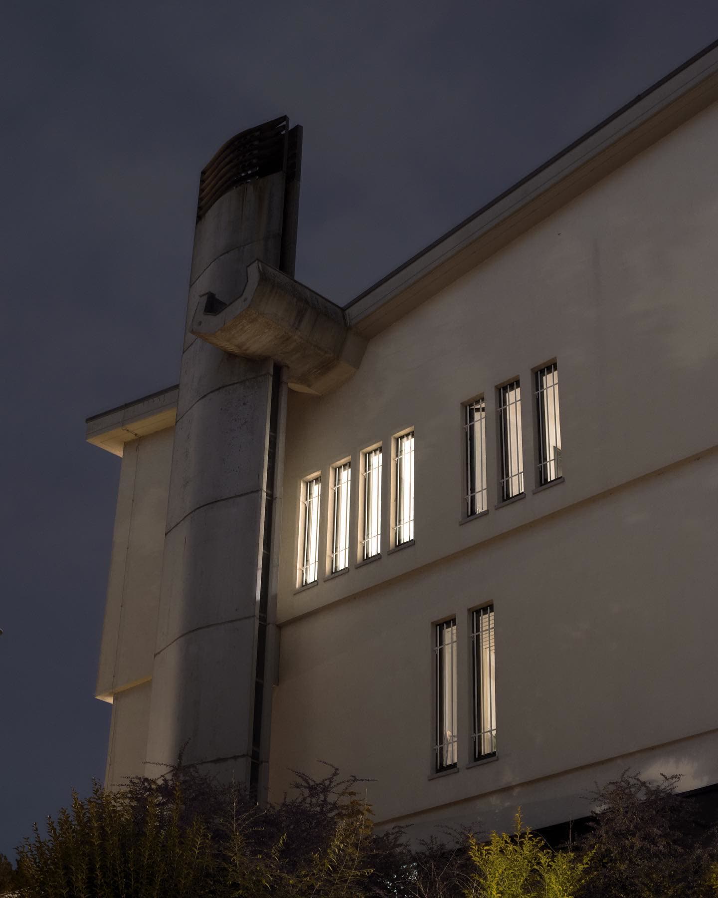 A modern building seen from below at night, a concrete chimney duct beside lit windows