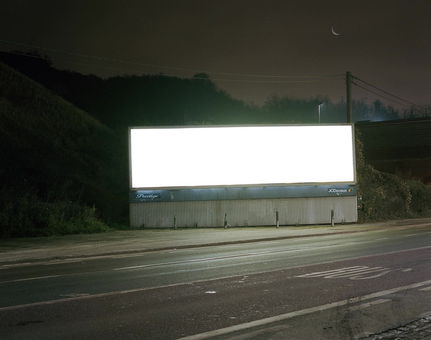 A blank billboard burns white against the night sky, a crescent moon rising above the trees