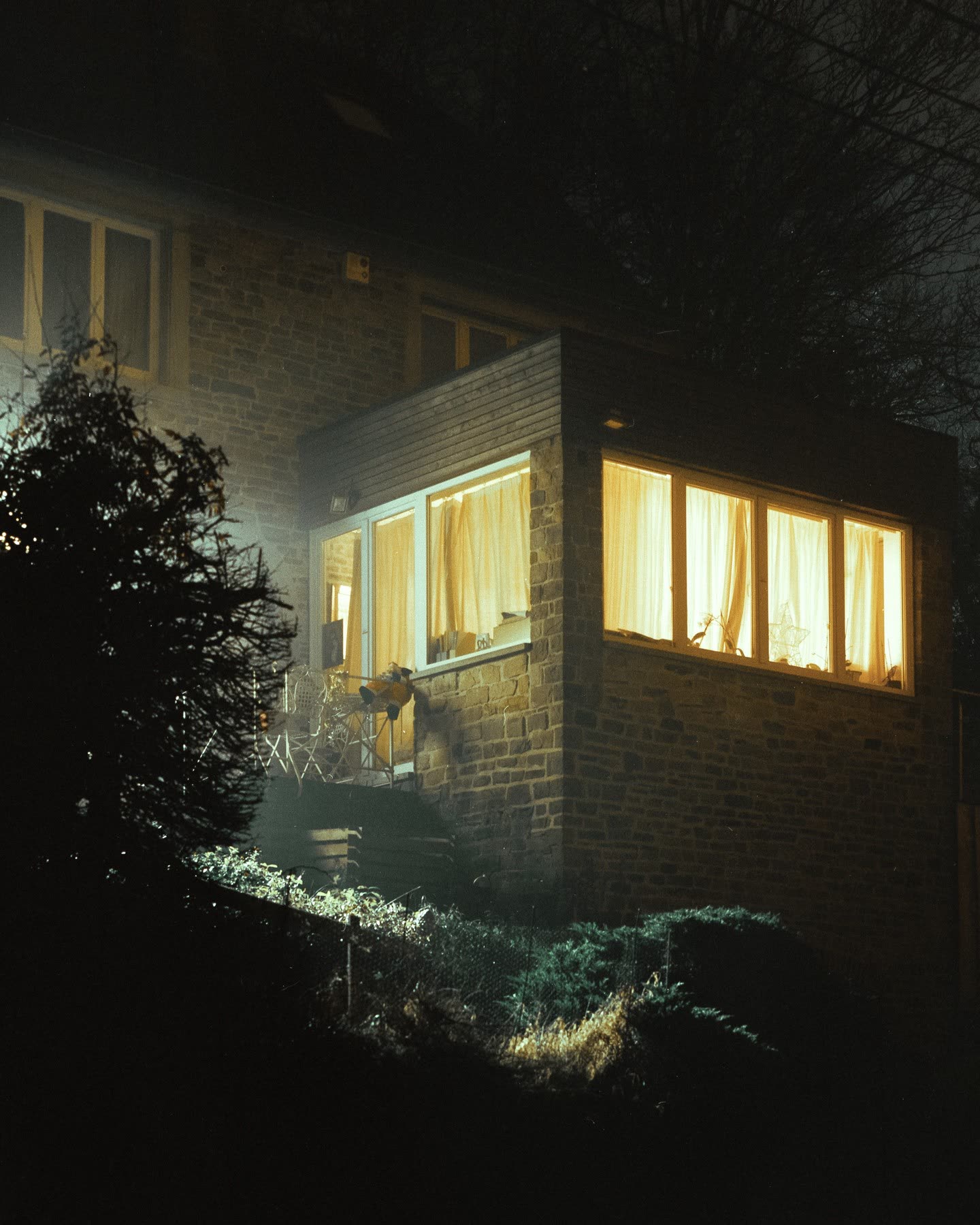 A brick house extension glowing warm yellow through curtained windows, dark trees around