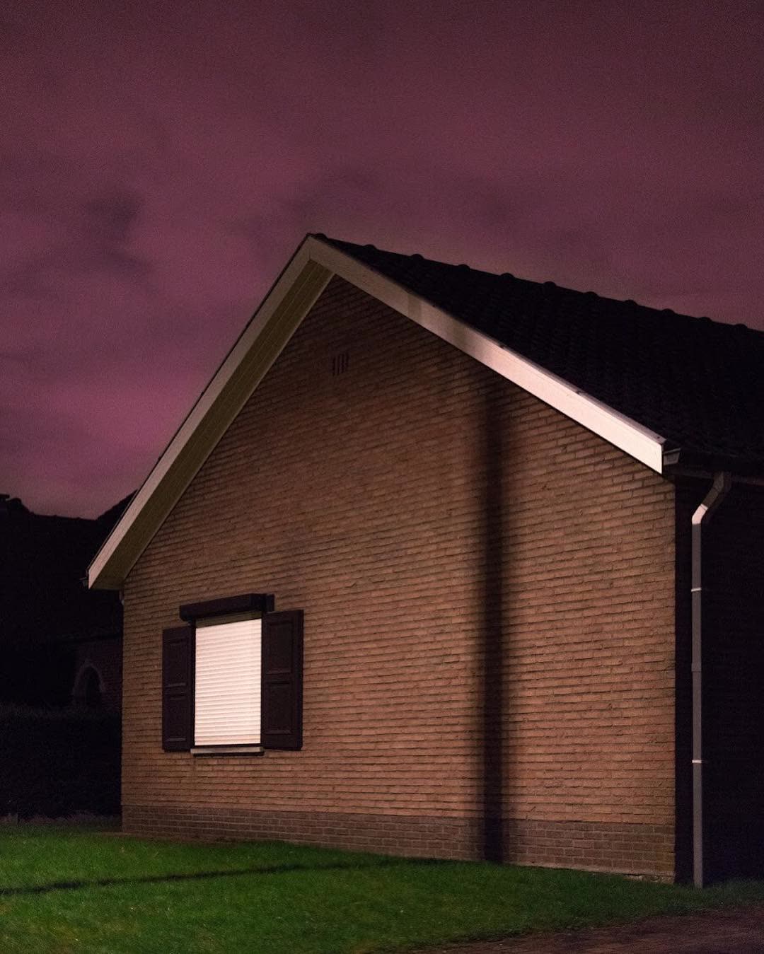 A brick house gable under a dramatic pink night sky, one shuttered window lit white