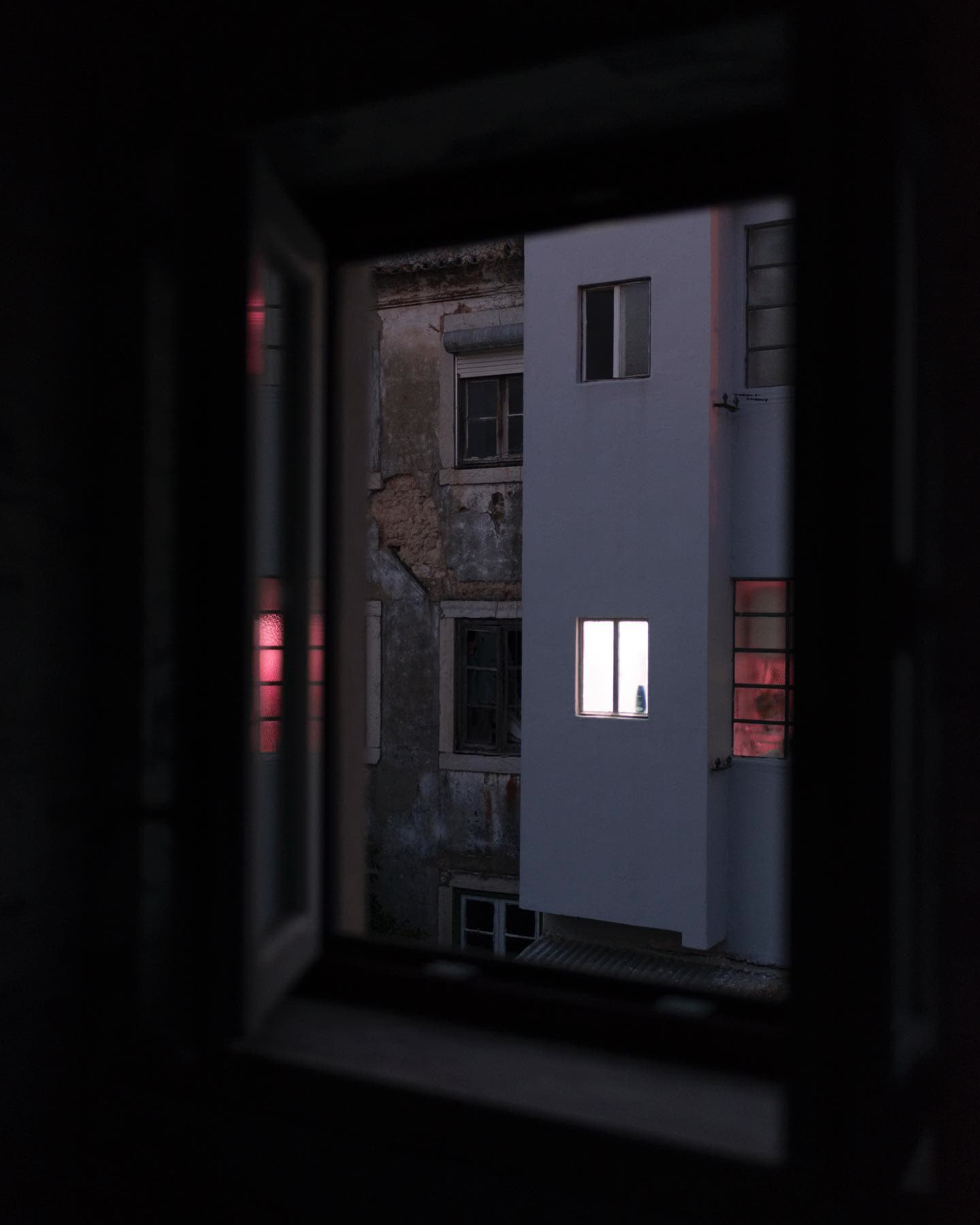 Looking through a dark window frame onto a white building lit by two glowing windows, pink reflections beyond